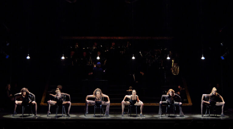 Six women on stage with band in the background.