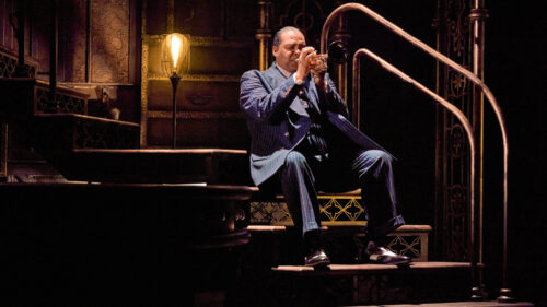 A man in a blue pin-striped suit sitting on a set of stairs, playing the trumpet.