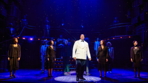 A man wearing a white suit jacket with black pants looking out into the audience. A spotlight is on him. 19 men and women dressed in formal wear surround him.