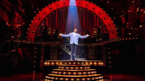 A man in a blue-checkered suit stands on an elevated stage with his arms spread, singing. A spotlight illuminates him.