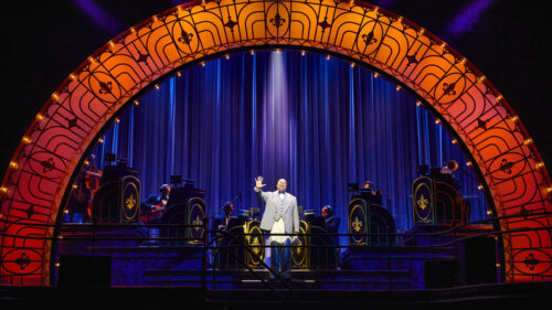 A man in a grey suit jacket with white pants stands under a gold-lit archway. A spotlight illuminates him and his right arm is stretched out in front of him, palm towards the camera.