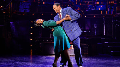 A man in a blue striped suit dips a women in a green dress while dancing. They are both smiling.