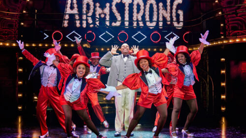 A man in a grey suit smiling with his hands next to his face, surrounded by seven dancers in matching red suits and top hats, posing.