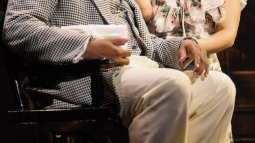 A man in a brown checkered suit sitting in a chair with a woman in a white flower dress kneeling down next to him touching his face, smiling.