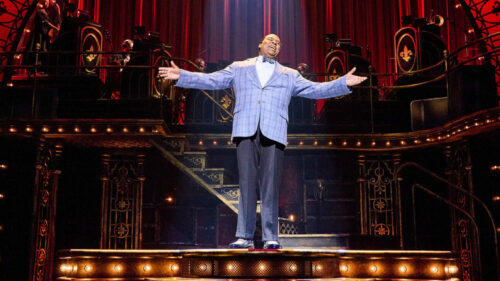 A man in a blue checkered suit standing on a stage with both of his arms spread, singing.